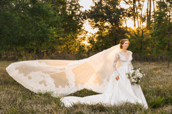 Bride in field with veil flowing behind her as sun peaks through the trees during Houston Bridal Portrait Session